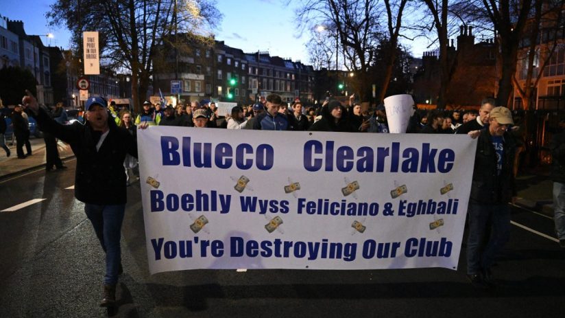 Football fans protest in the street holding a banner: Blueco Clearlake Boehly Wyss Feliciano & Eghbali Youre Destroying Ou...
