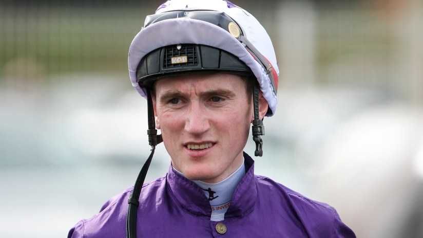 Jockey Jack Kennedy in purple silks and white helmet with KEP Italia branding, looking forward at a racecourse.
