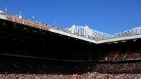 Football match in a stadium, Sir Alex Ferguson Stand visible on the roof, with fans and players on the field.