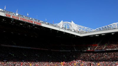 Football match in a stadium, Sir Alex Ferguson Stand visible on the roof, with fans and players on the field.