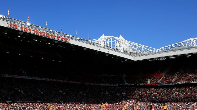 Football match in a stadium, Sir Alex Ferguson Stand visible on the roof, with fans and players on the field.