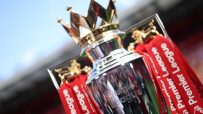 Close-up of the shining Premier League trophy with red ribbons, set against a blurred stadium background.