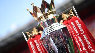 Close-up of the shining Premier League trophy with red ribbons, set against a blurred stadium background.