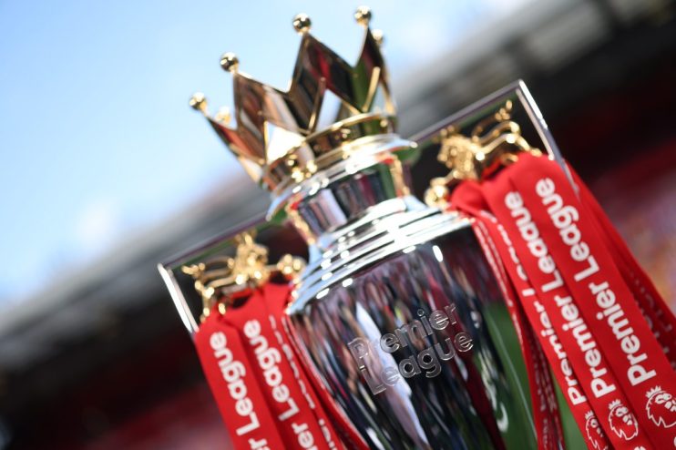 Close-up of the shining Premier League trophy with red ribbons, set against a blurred stadium background.