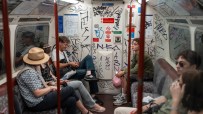 Passengers inside a graffiti-covered London Underground train car, some looking at phones, others observing the interior.