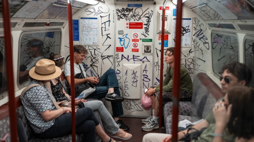 Passengers inside a graffiti-covered London Underground train car, some looking at phones, others observing the interior.