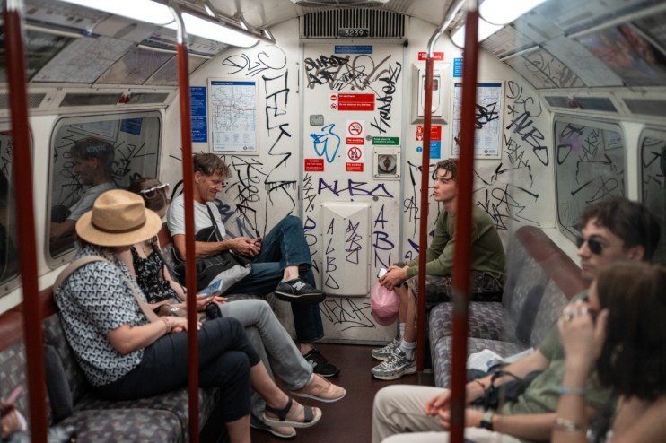 Passengers inside a graffiti-covered London Underground train car, some looking at phones, others observing the interior.
