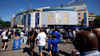 Crowds of fans gather outside the Chelsea FC stadium on a sunny day, with a Matchday Programmes On Sale sign visible.