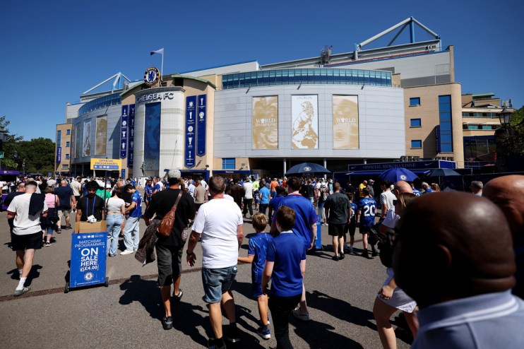Crowds of fans gather outside the Chelsea FC stadium on a sunny day, with a Matchday Programmes On Sale sign visible.