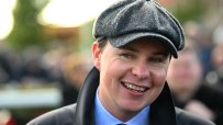 Smiling man in a tweed flat cap, blue shirt, and dark jacket, looking forward.