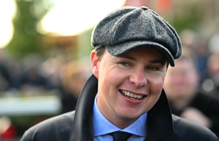 Smiling man in a tweed flat cap, blue shirt, and dark jacket, looking forward.