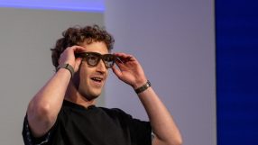 Mark Zuckerberg smiling while adjusting smart glasses on his face, wearing a black shirt and two wristbands