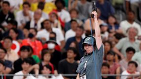 Female official in headset and hat raising a starting pistol at a sporting event, crowd blurred in background