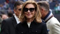 Smiling woman with blonde hair and black sunglasses in a black coat