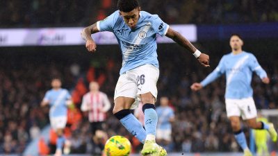 A male footballer in a light blue Manchester City kit, number 26, kicks a yellow soccer ball on a green field.