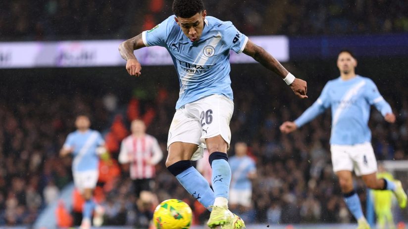 A male footballer in a light blue Manchester City kit, number 26, kicks a yellow soccer ball on a green field.