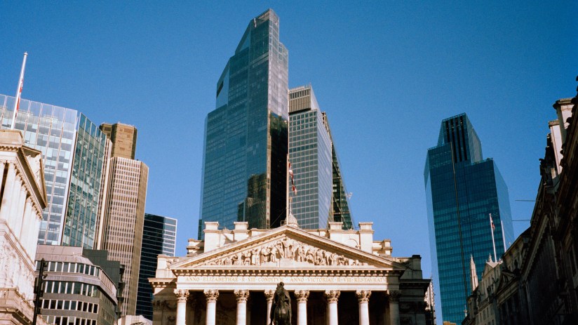 Historic Royal Exchange building in London with modern skyscrapers behind, clear blue sky.