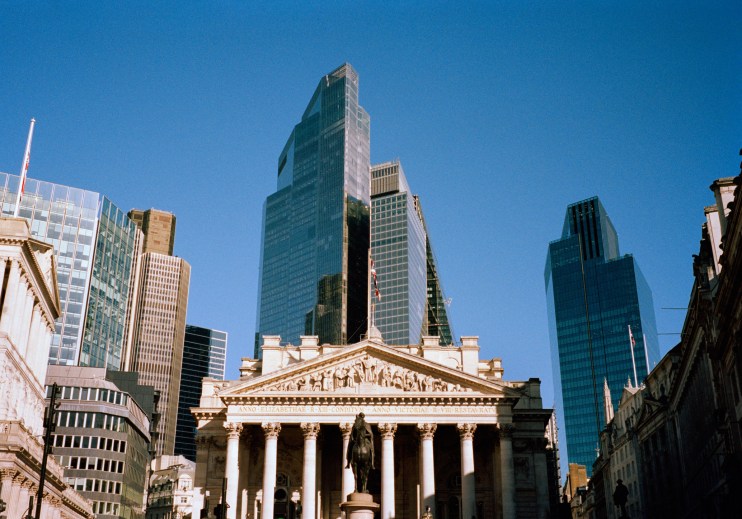 Historic Royal Exchange building in London with modern skyscrapers behind, clear blue sky.