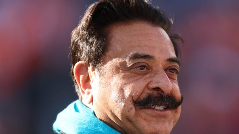 Shahid Khan, businessman, smiling and wearing a bright blue scarf, looking to the right with a blurred background.