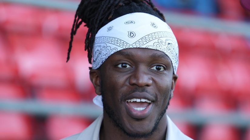 KSI with dreadlocks and a white bandana, smiling with an open mouth, red stadium seats blurred in background.