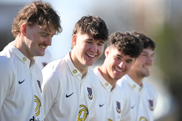 Four young men in white collared shirts with logos, smiling and looking down, outdoors.