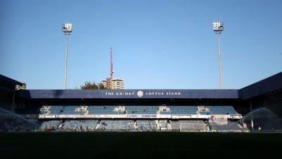 Loftus Road stadium with THE GO-OUT LOFTUS STAND sign, floodlights, and sprinklers on the pitch.
