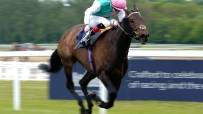 Jockey in pink helmet and green/white silks riding a brown racehorse at full gallop on a green track.