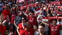 Liverpool FC fans cheering in a stadium, holding up red scarves and wearing team jerseys.