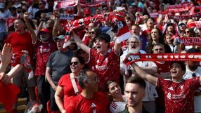 Liverpool FC fans cheering in a stadium, holding up red scarves and wearing team jerseys.