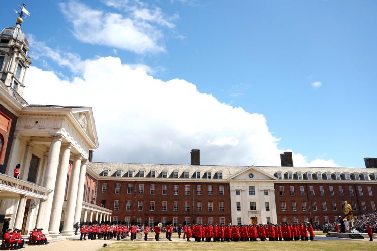 British soldiers in red uniforms parade in front of historic buildings under a partly cloudy sky.