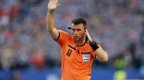 Soccer referee in an orange FIFA uniform with a headset, raising his hand on the field during a match