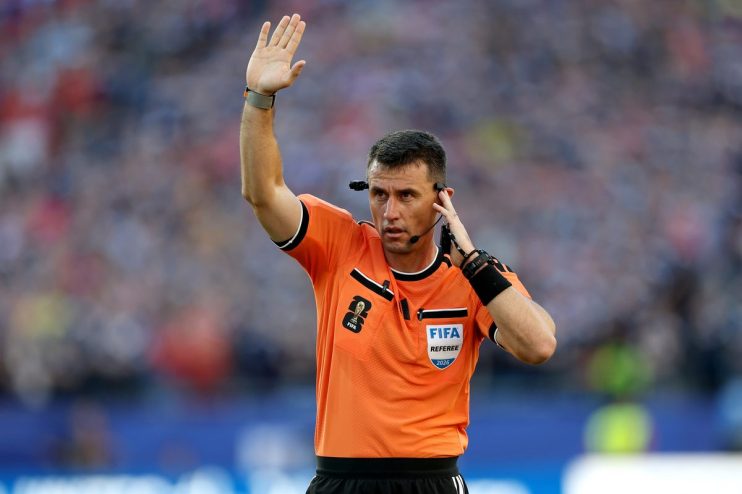 Soccer referee in an orange FIFA uniform with a headset, raising his hand on the field during a match
