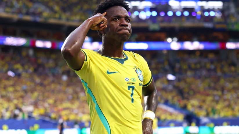 Vinicius Jr. celebrates a goal in a yellow Brazil jersey during a football match, with stadium lights in background.