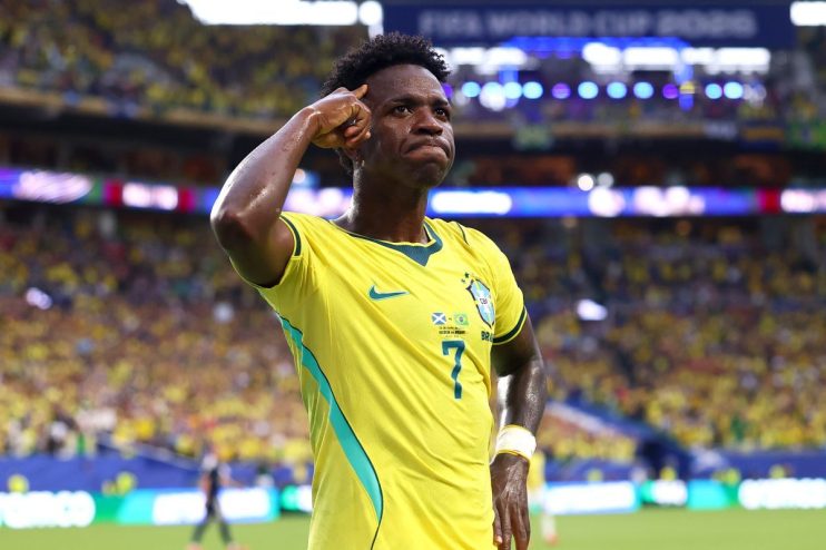 Vinicius Jr. celebrates a goal in a yellow Brazil jersey during a football match, with stadium lights in background.
