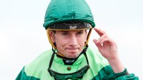 Jockey in green and light green silks, wearing a helmet and goggles, adjusting his headwear.
