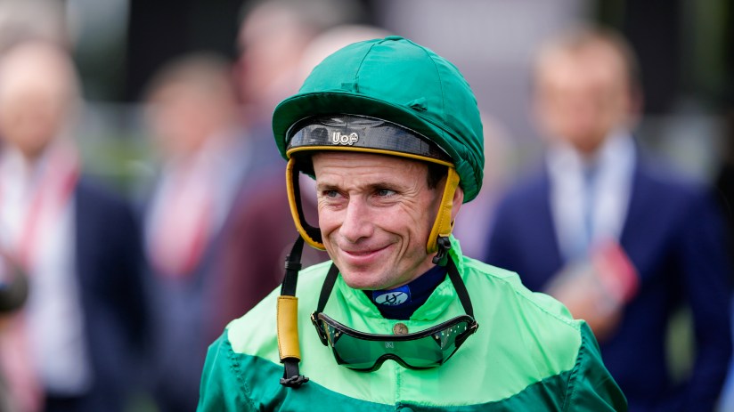 Jockey in green and dark green silks, wearing a green helmet, smiling with goggles around his neck