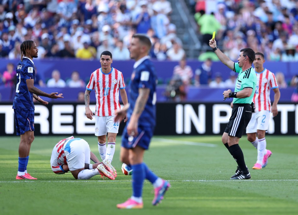 Soccer referee issuing a yellow card to a player during a match, with other players and spectators in background.