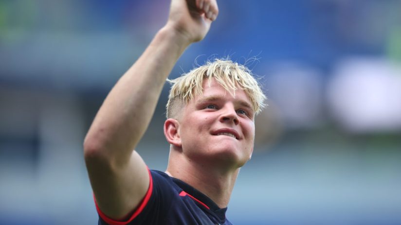 A young man with blonde hair and a dark shirt, clenching his fist and smiling, likely celebrating or cheering.