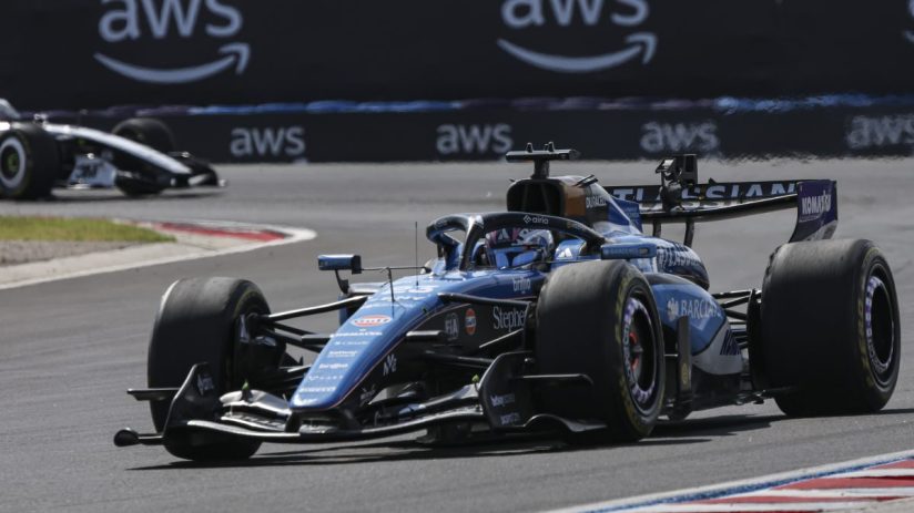Blue Formula 1 race car on track with AWS branding on the barrier, another car in the background.