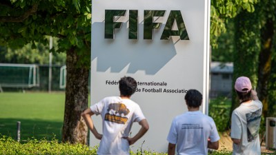 Three people view the FIFA sign and official name in front of a green soccer field.