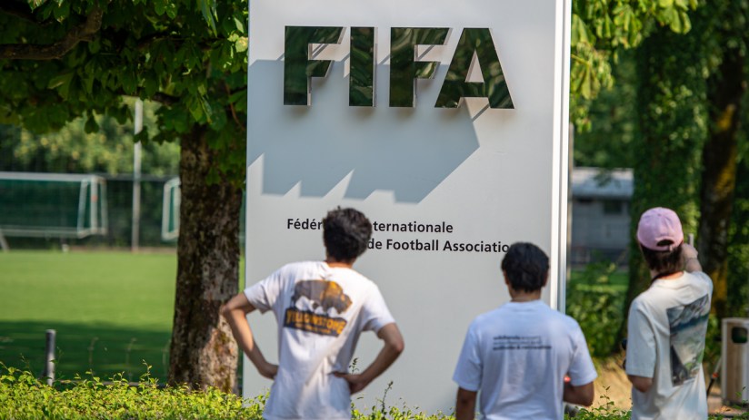 Three people view the FIFA sign and official name in front of a green soccer field.