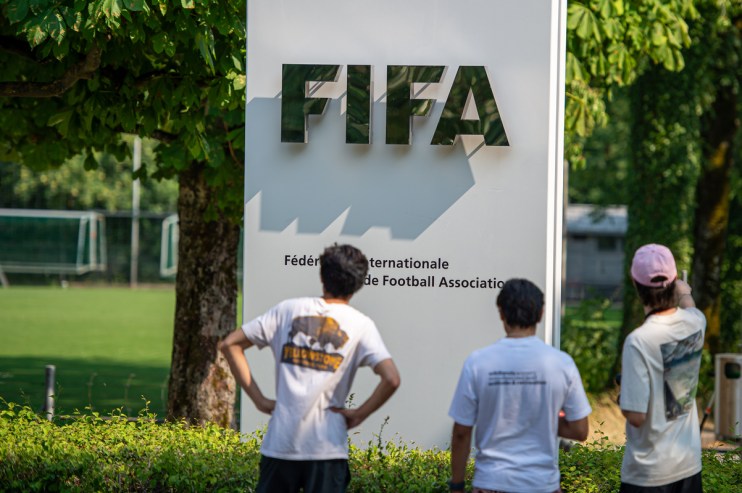Three people view the FIFA sign and official name in front of a green soccer field.