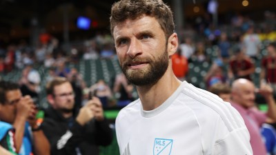 Thomas M&uuml;ller in a white MLS jersey looking forward with fans in the background at a stadium event.