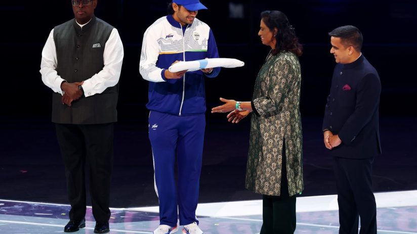 Indian athlete in blue tracksuit receives a white scroll from a woman in a green traditional dress, flanked by two men in ...