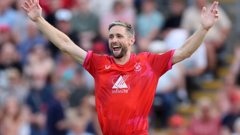 Chris Woakes, cricketer in a red uniform, raising his arms in celebration on the field.