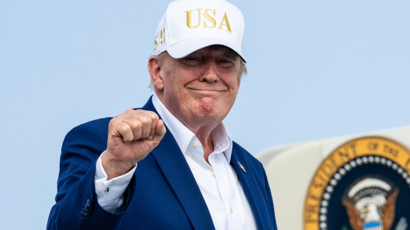 Donald Trump in a white USA hat, blue suit, and white shirt, making a fist, with a presidential seal visible