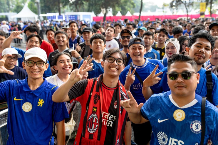 Enthusiastic football fans, some in Chelsea and AC Milan jerseys, smiling and making peace signs at an event.