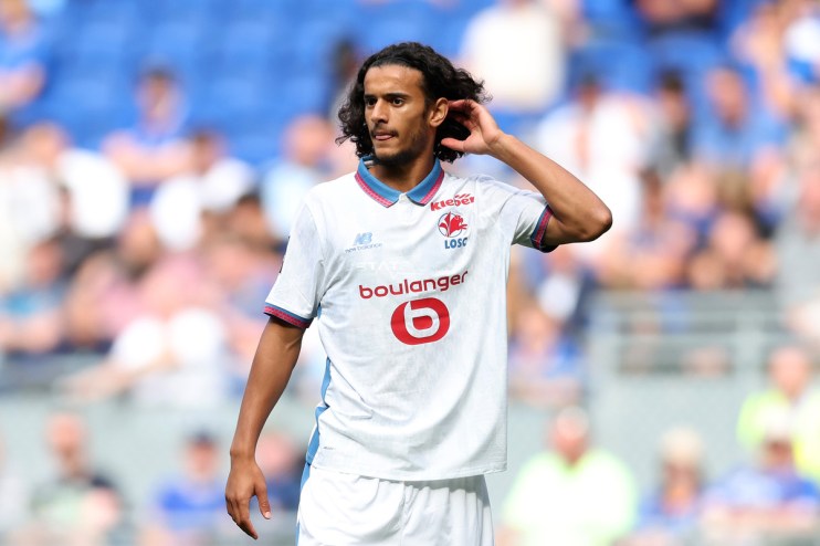 A male soccer player with long dark curly hair in a white LOSC Lille jersey adjusts his hair on the field.