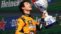 Lando Norris, in an orange racing suit, holds up a white and blue patterned trophy, cheering on a podium.