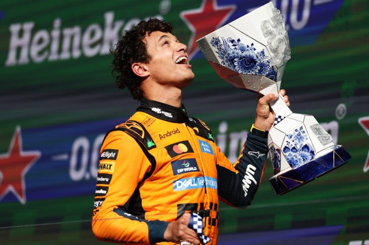 Lando Norris, in an orange racing suit, holds up a white and blue patterned trophy, cheering on a podium.